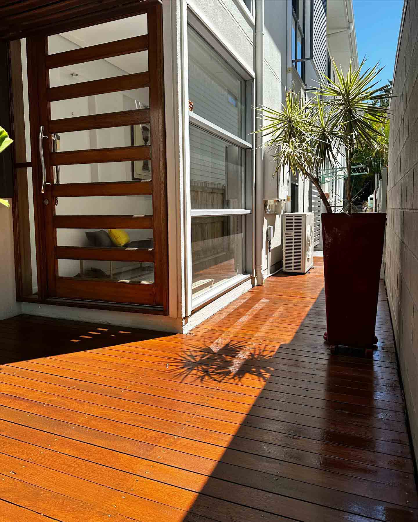 Sunlit oiled timber side deck by a timber front door and red pot plant