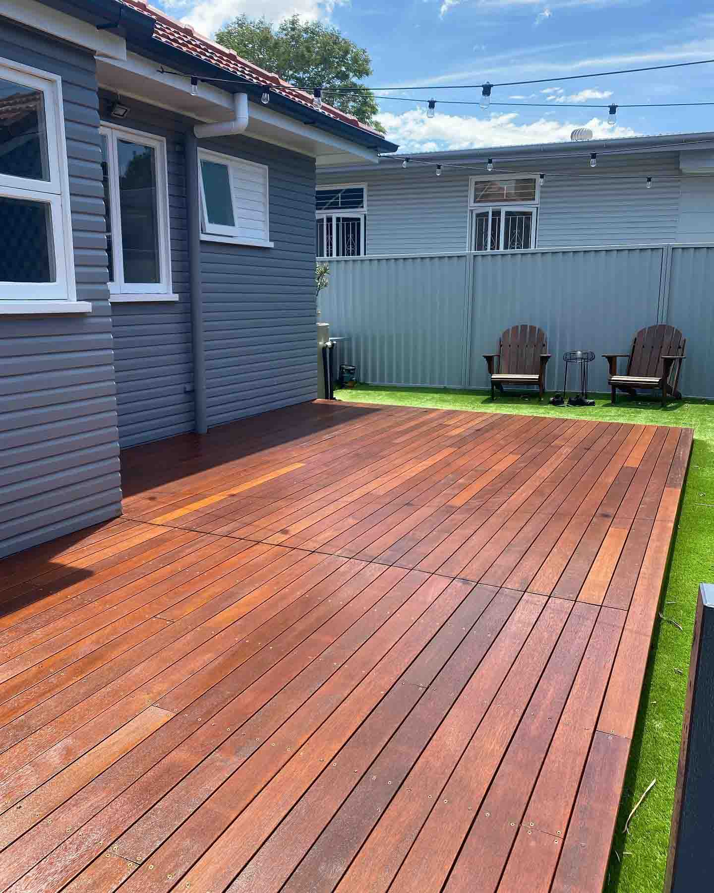Oiled timber deck beside a grey weatherboard house and artificial turf, with two chairs