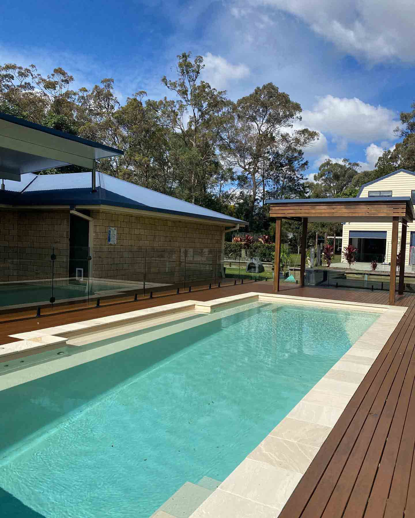 Timber deck along a rectangular pool with a cabana and trees behind
