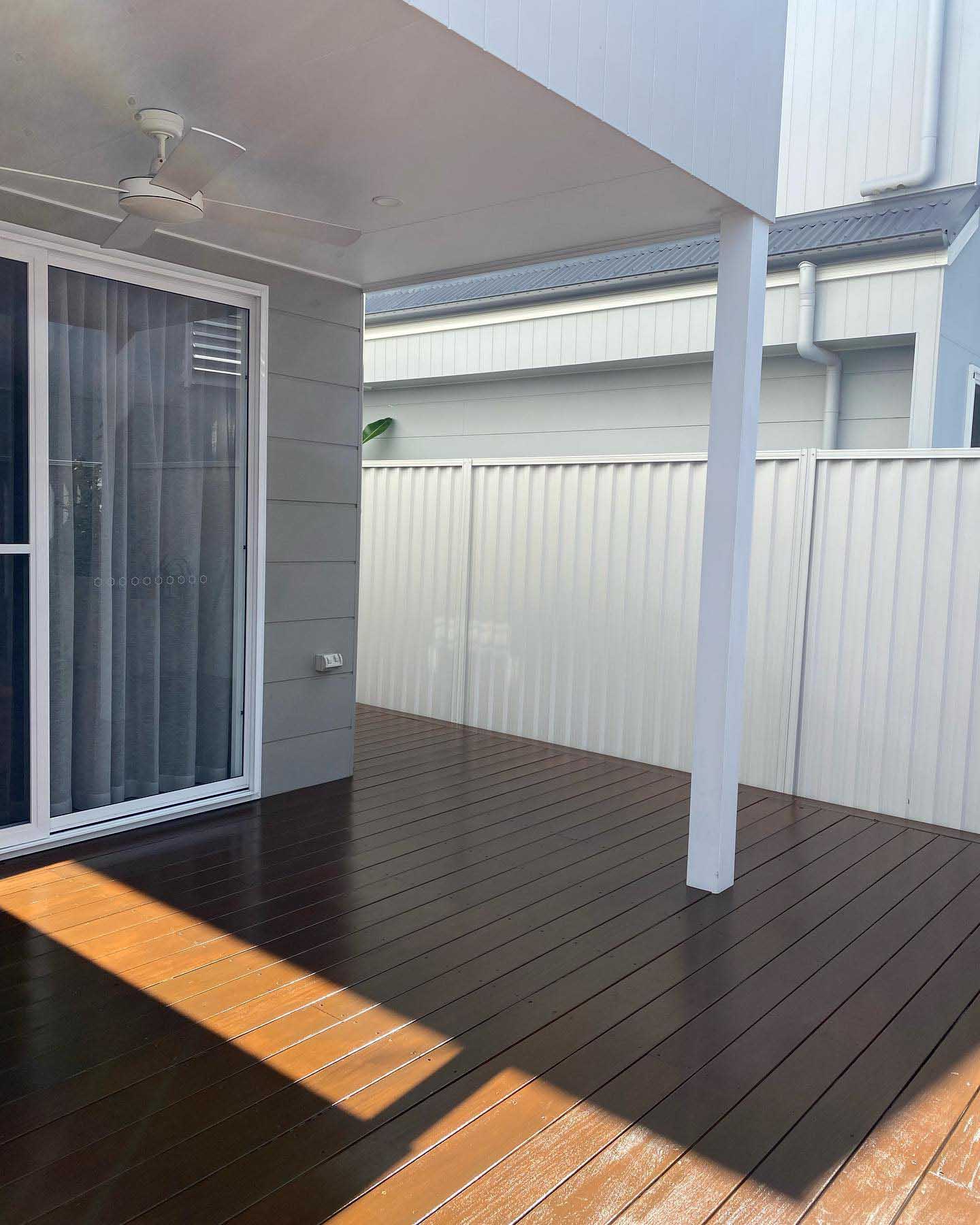 Dark brown-coated deck under a patio beside sliding doors and a pale fence