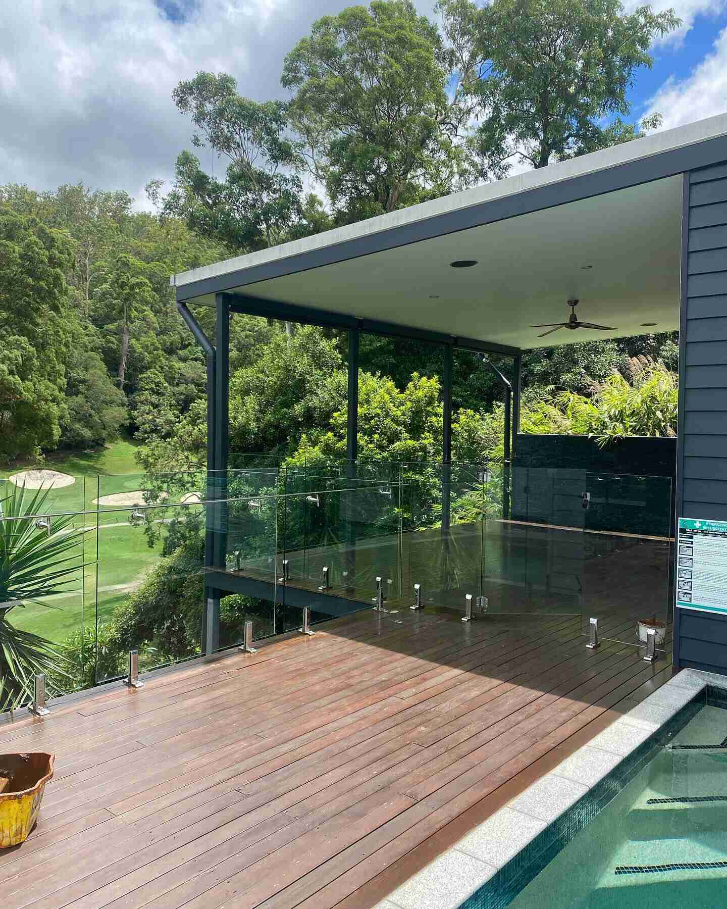 Covered timber deck with glass balustrade beside a pool, overlooking a golf course