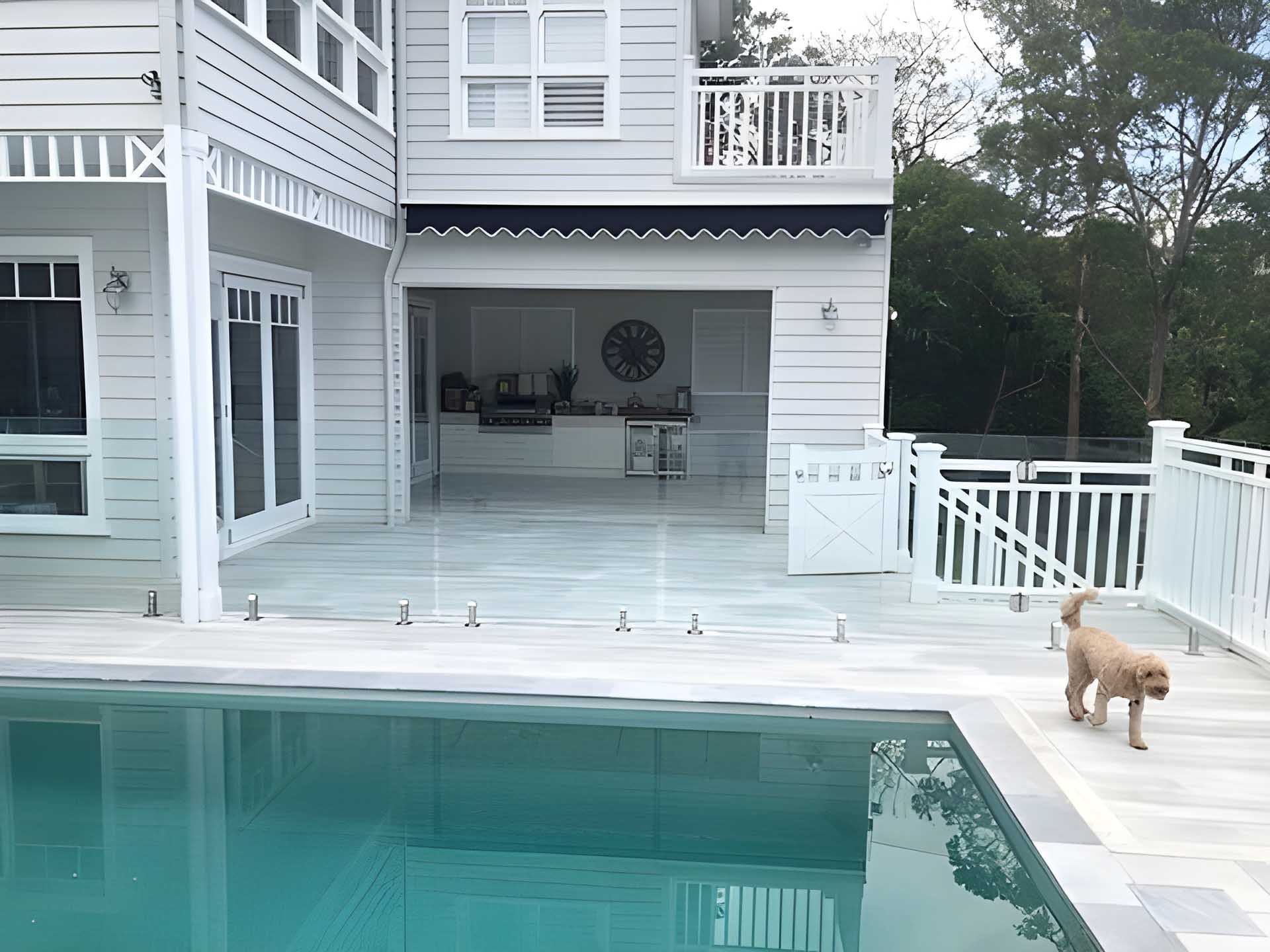 Pale grey deck around a pool beside a white two-storey house, with a small dog
