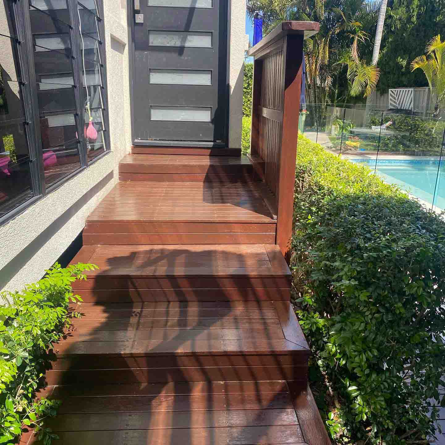 Oiled timber steps to a dark front door beside a hedge and pool