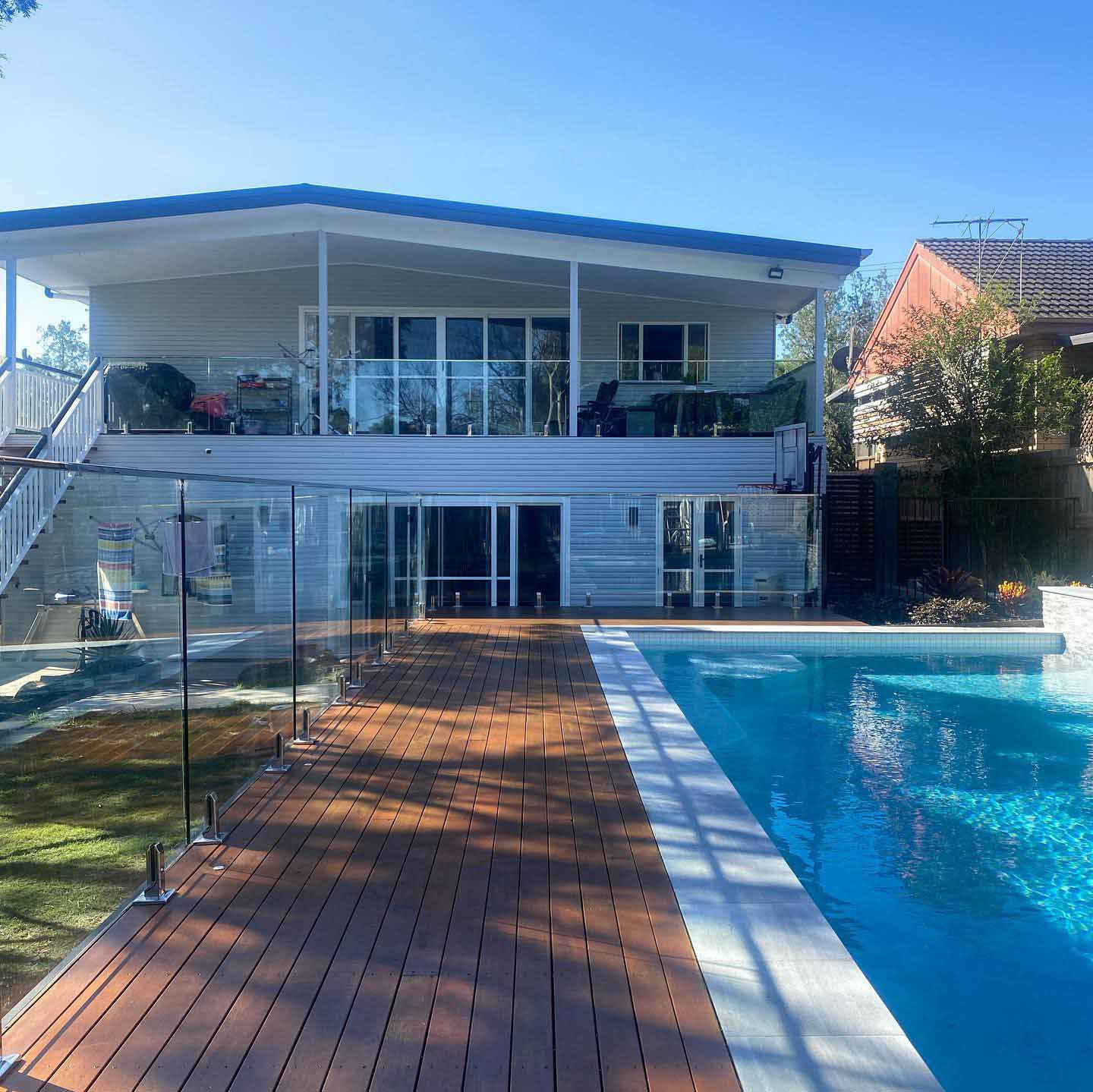 Long timber deck beside a pool with glass fencing below a two-storey house