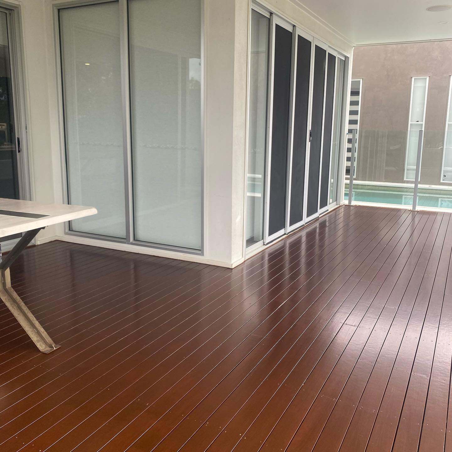 Glossy oiled timber deck under a patio beside sliding doors, pool behind