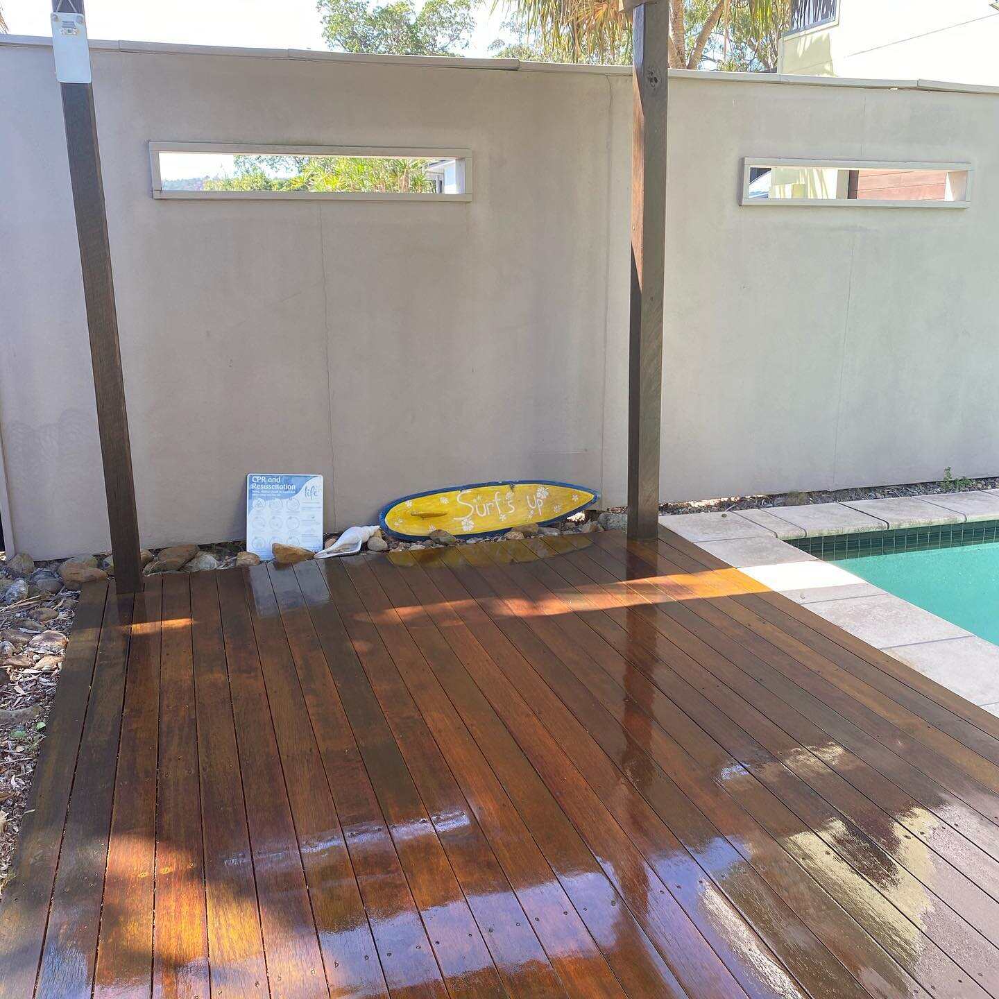 Glossy wet oiled timber deck by a rendered wall and pool, with a surfboard