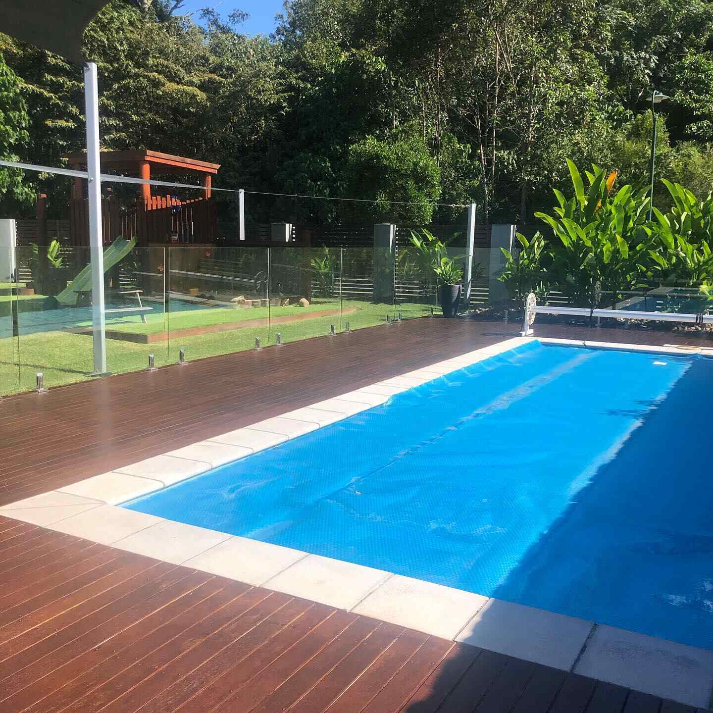 Oiled timber deck around a pool with a blue cover and glass fencing