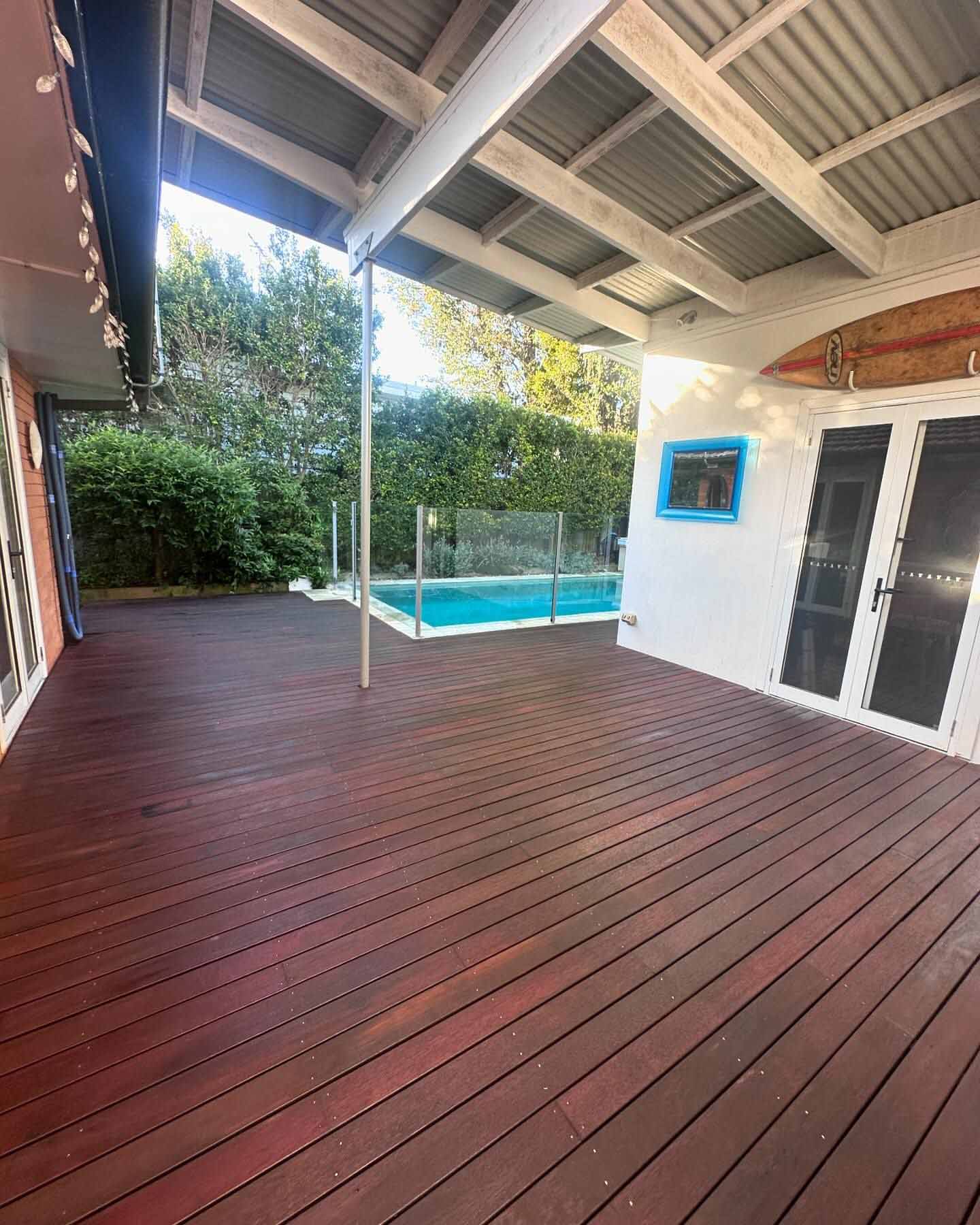 Dark red oiled timber deck under a patio roof beside a fenced pool