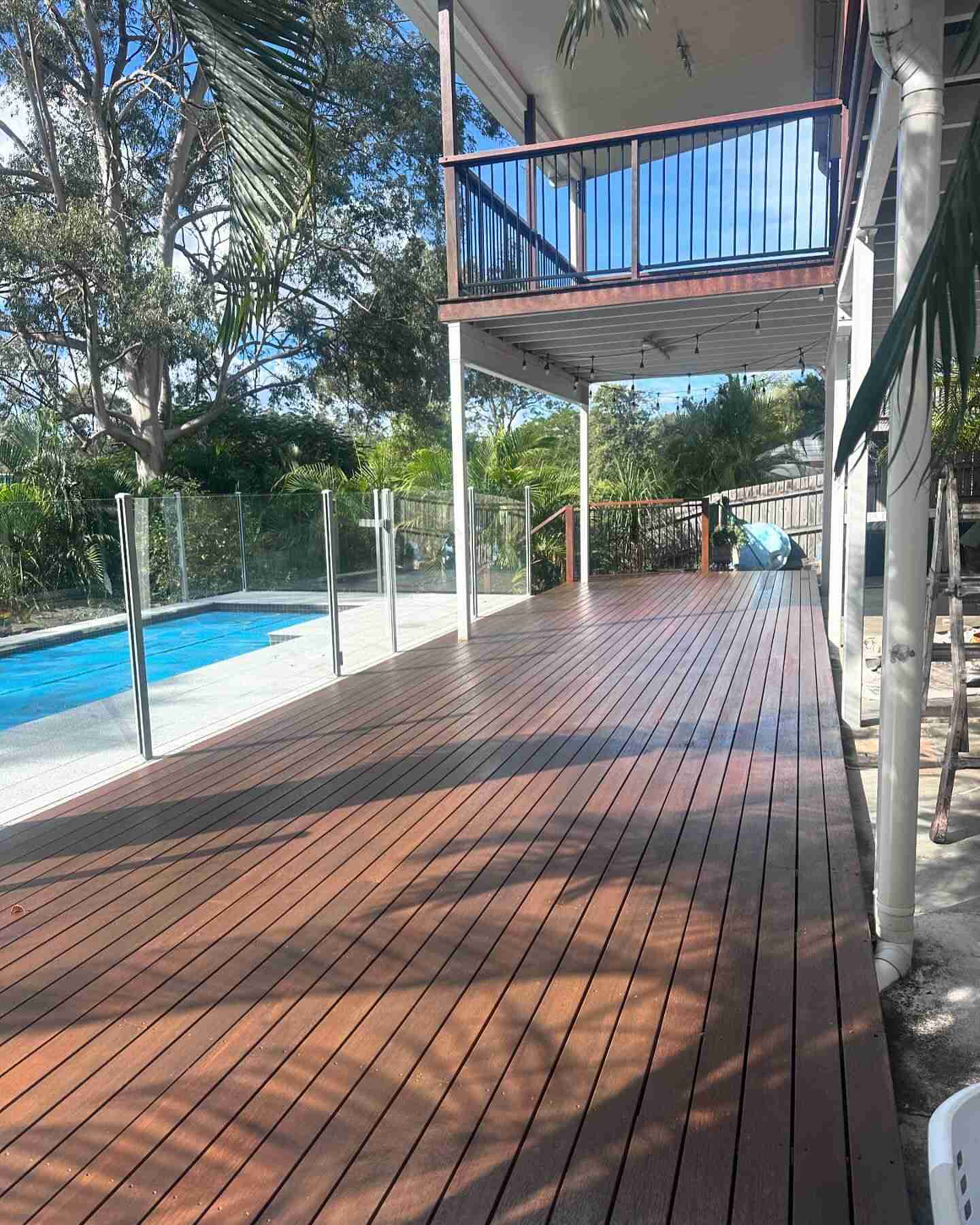 Oiled timber deck along a glass-fenced pool beneath an upper balcony