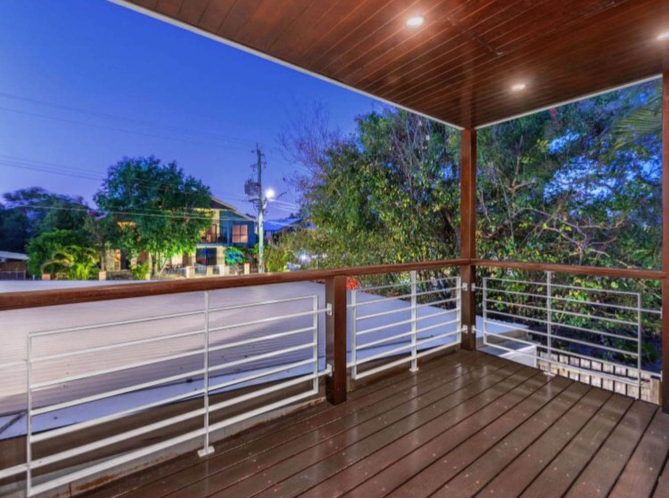 Oiled timber balcony with white balustrade under a timber-lined ceiling at dusk